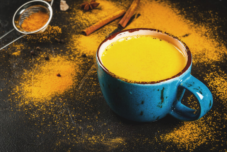 A blue mug filled with golden-yellow liquid sits on a dark surface sprinkled with turmeric powder and surrounded by cinnamon sticks and a small strainer.