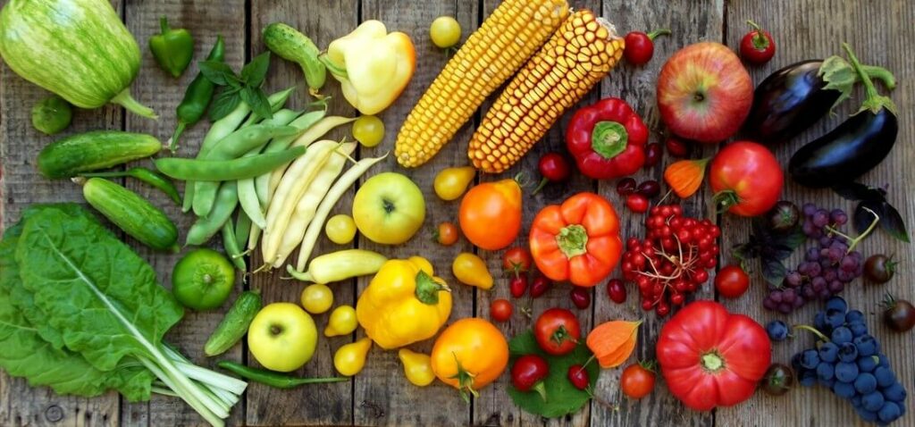 Assorted fresh vegetables, fruits, and grains arranged on a wooden surface, including cucumbers, peppers, corn, leafy greens, tomatoes, eggplants, apples, berries, and legumes.