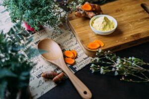 A wooden spoon rests on turmeric powder with fresh turmeric root, carrot slices, and dried herbs arranged on a wooden board surrounded by potted herbs and greenery.