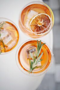Three glasses of amber cocktails garnished with grapefruit slices and fresh rosemary sprigs on a white surface.