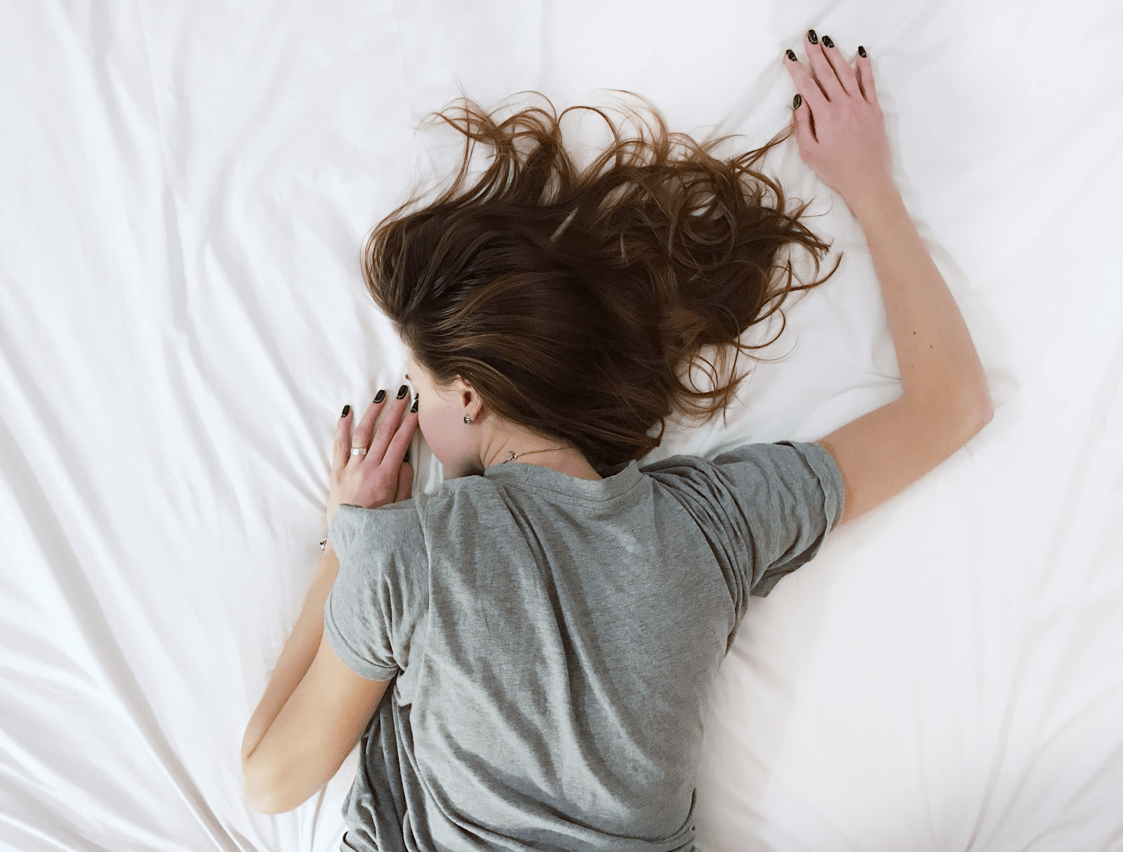 Woman with brown hair sleeping face-down on white bedding wearing a gray t-shirt.