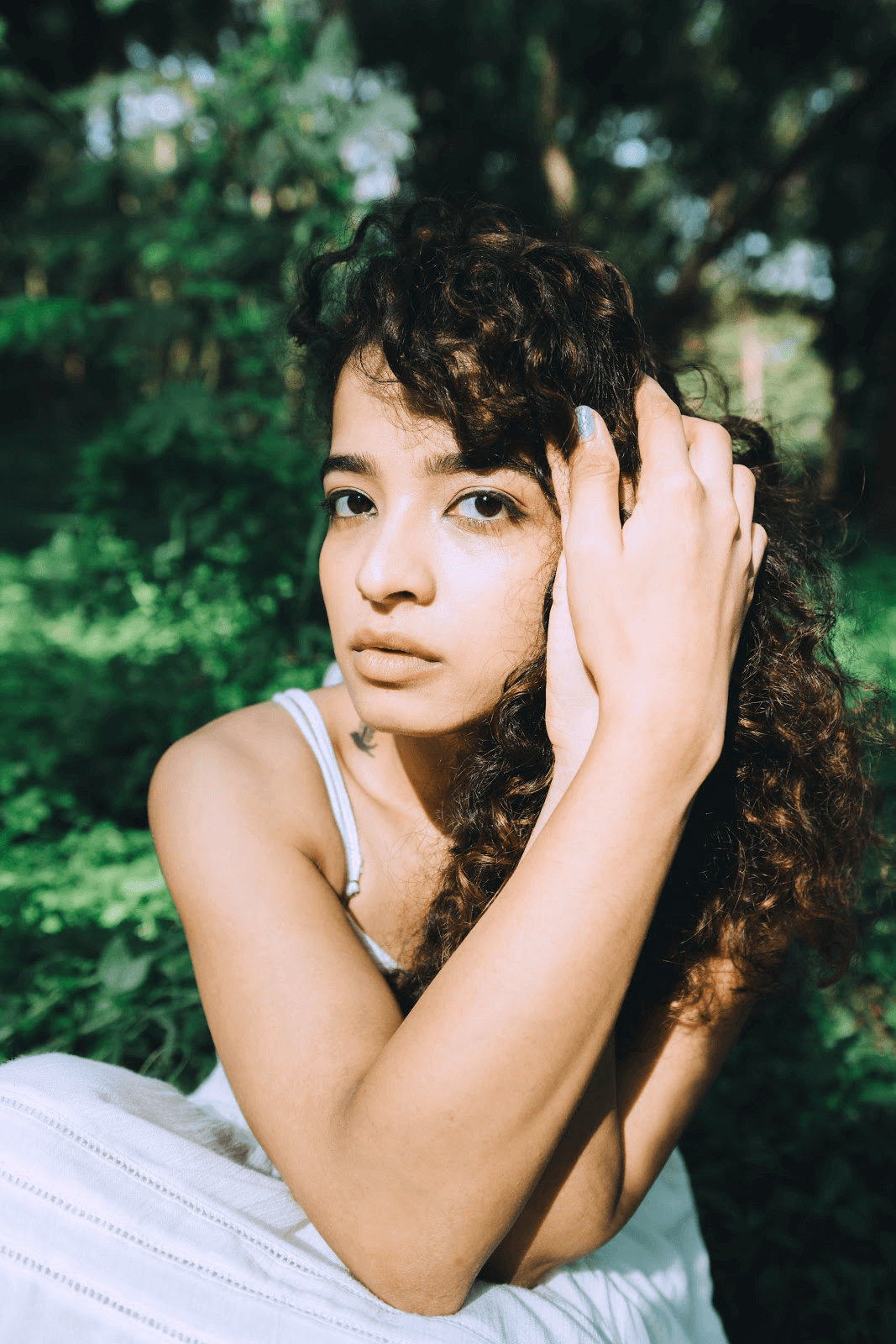 A person with curly dark hair poses thoughtfully with their hand near their face against a lush green garden background.