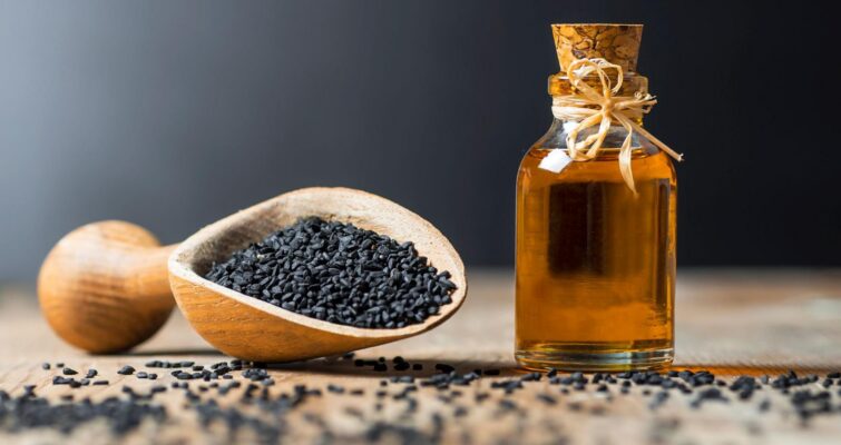 A wooden scoop full of black seeds near a small bottle of black seed oil