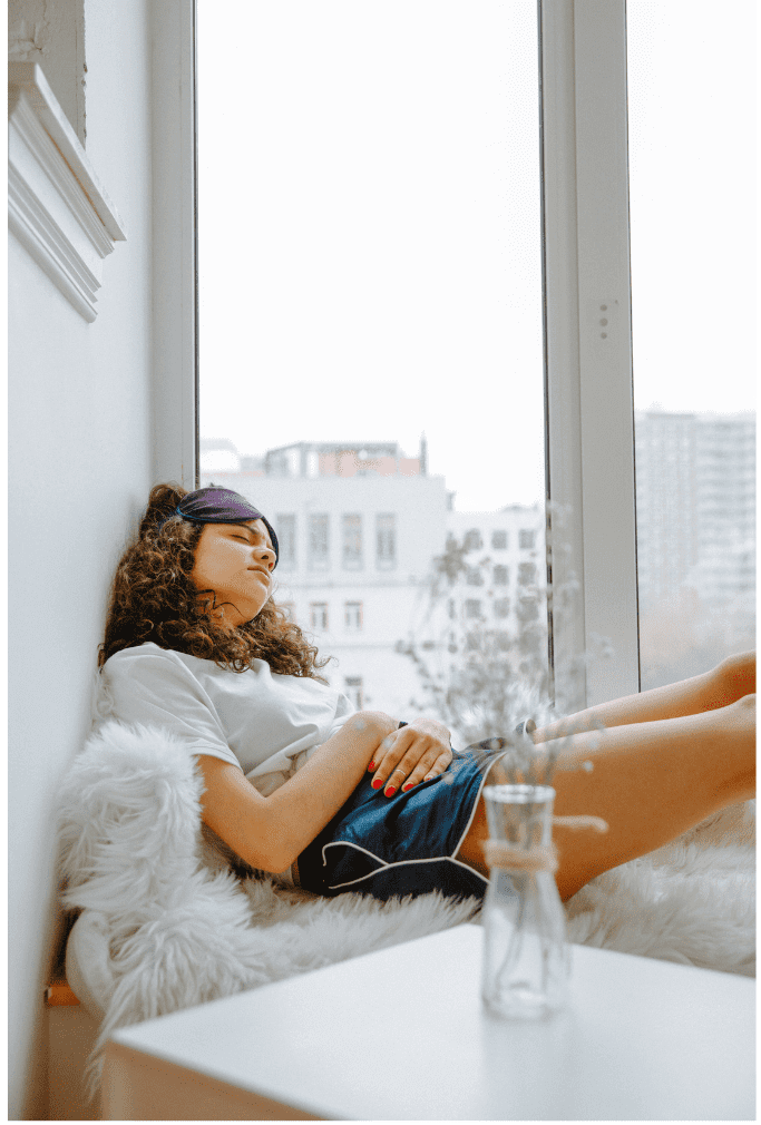 A woman with curly hair and a purple headband relaxes by a large window in a high-rise apartment, sitting on a white furry seat with her legs extended while gazing outward at the city skyline.
