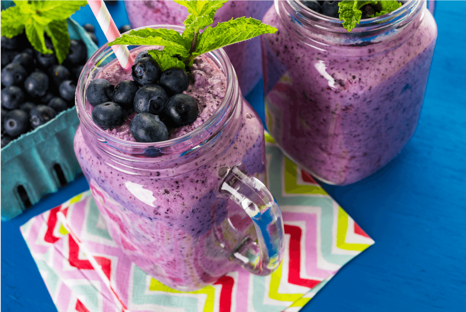Two purple blueberry smoothies garnished with fresh mint leaves and topped with blueberries, served in glass jars with striped straws on a colorful patterned napkin against a bright blue background.