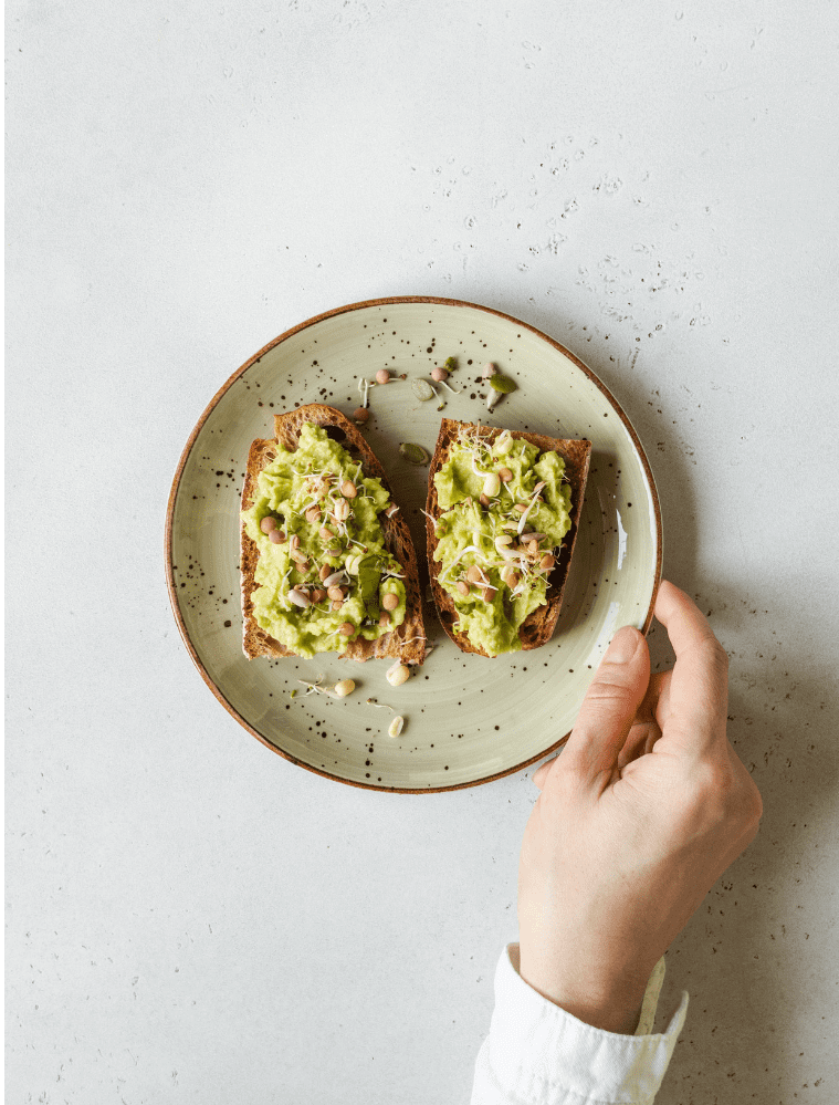 A hand holds a speckled ceramic plate with two slices of toasted bread topped with mashed avocado and chopped nuts.