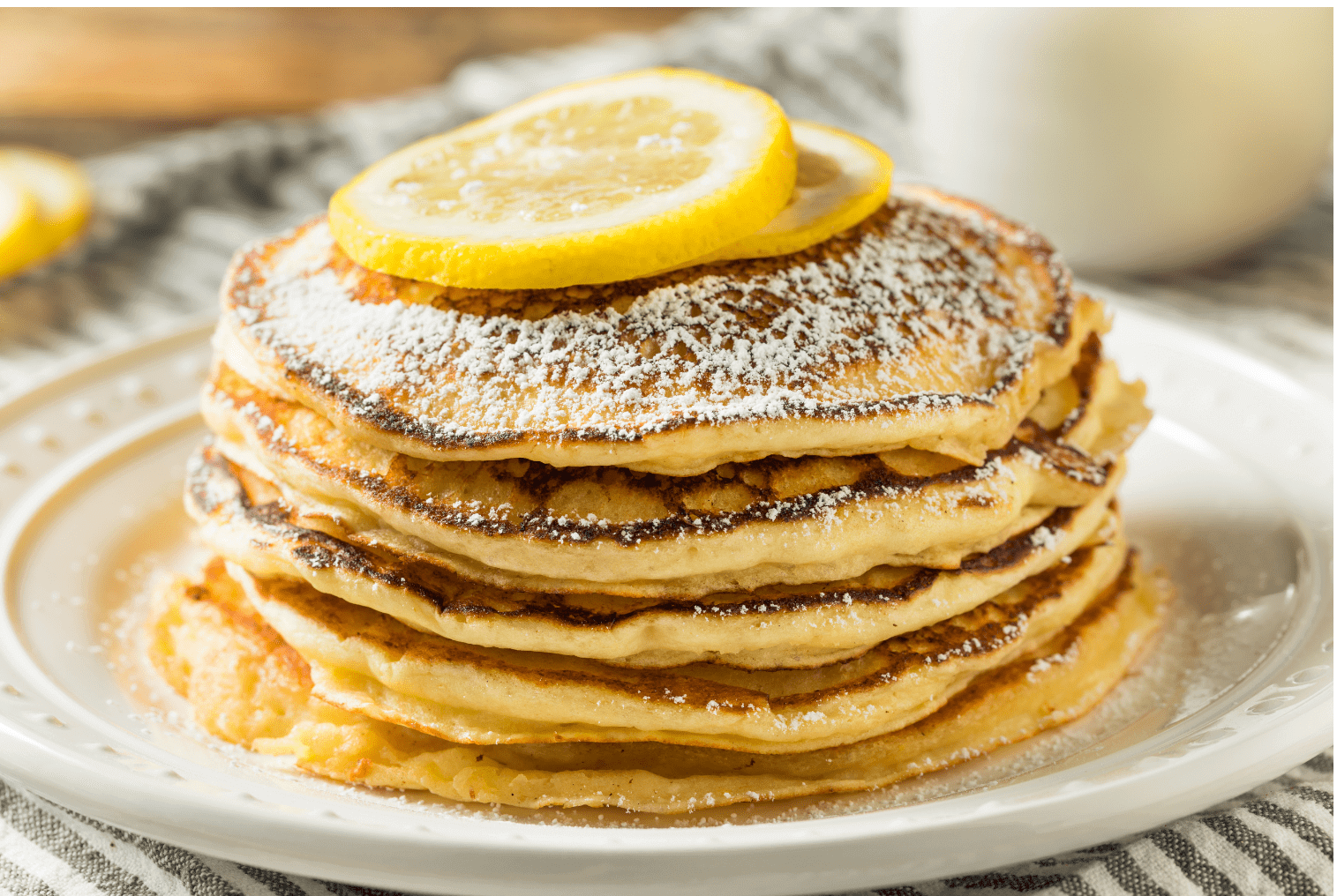 Stack of five fluffy pancakes dusted with powdered sugar and topped with a fresh lemon slice on a white plate.