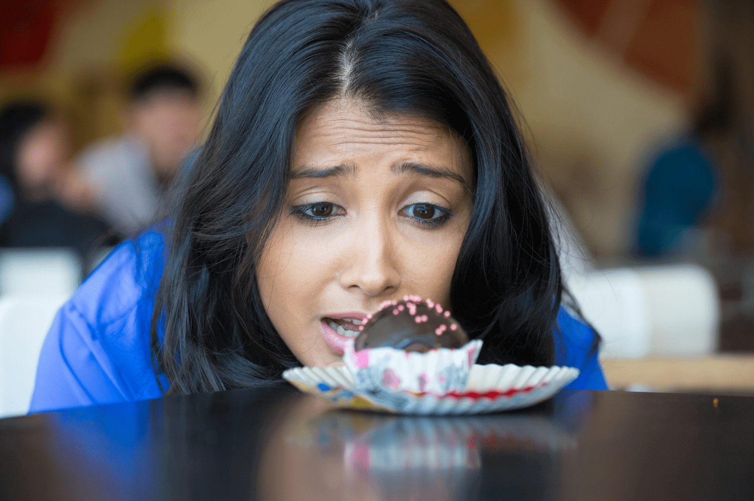 A woman in a blue shirt leans toward a plate with a chocolate cupcake, looking at it with a concerned or uncertain expression.
