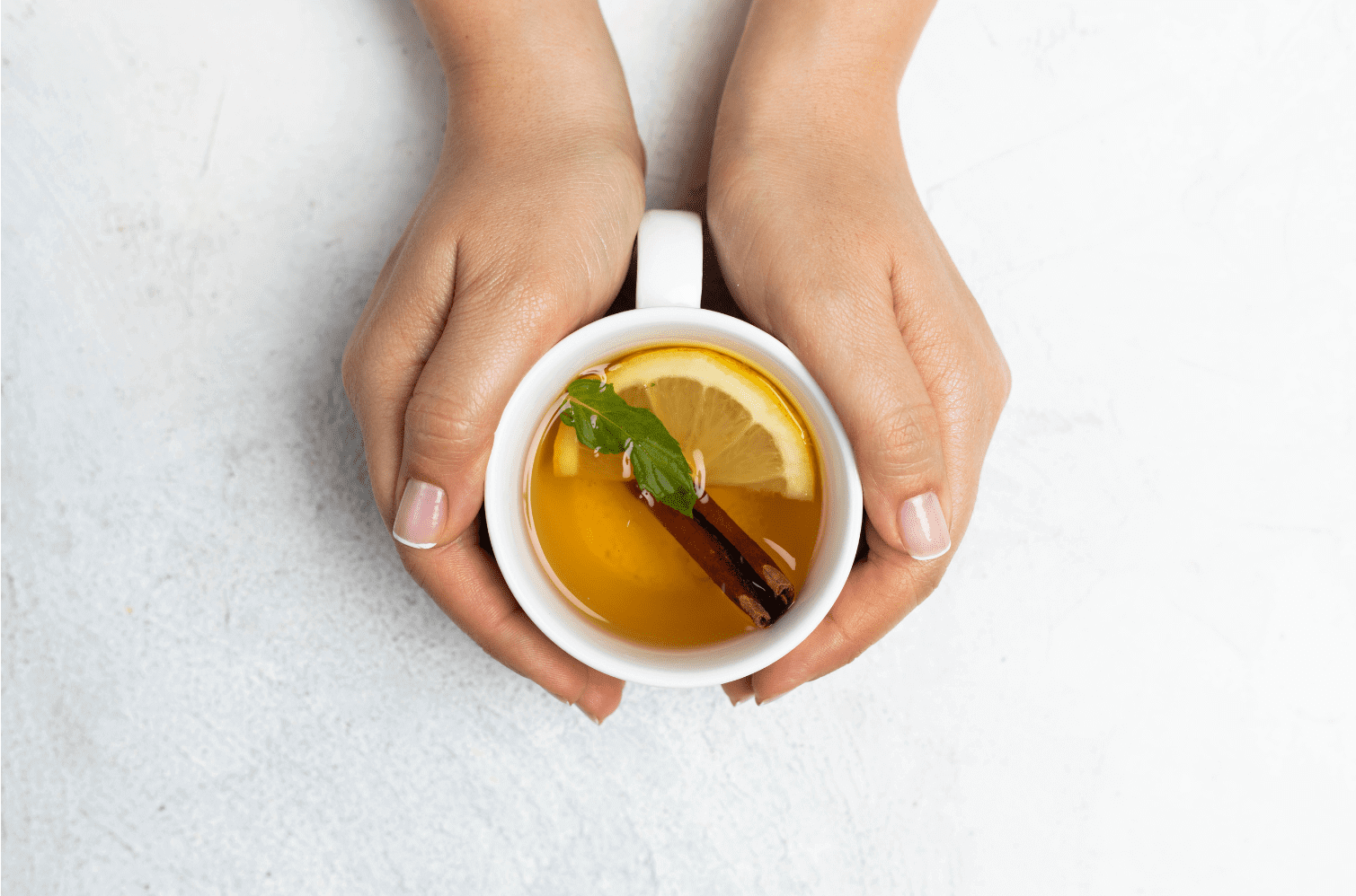 Hands cradling a white mug of herbal tea with lemon slice, cinnamon stick, and fresh mint leaf.