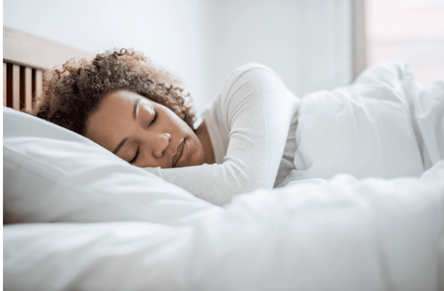 A person with curly brown hair sleeps peacefully on white bedding with eyes closed and a relaxed facial expression.