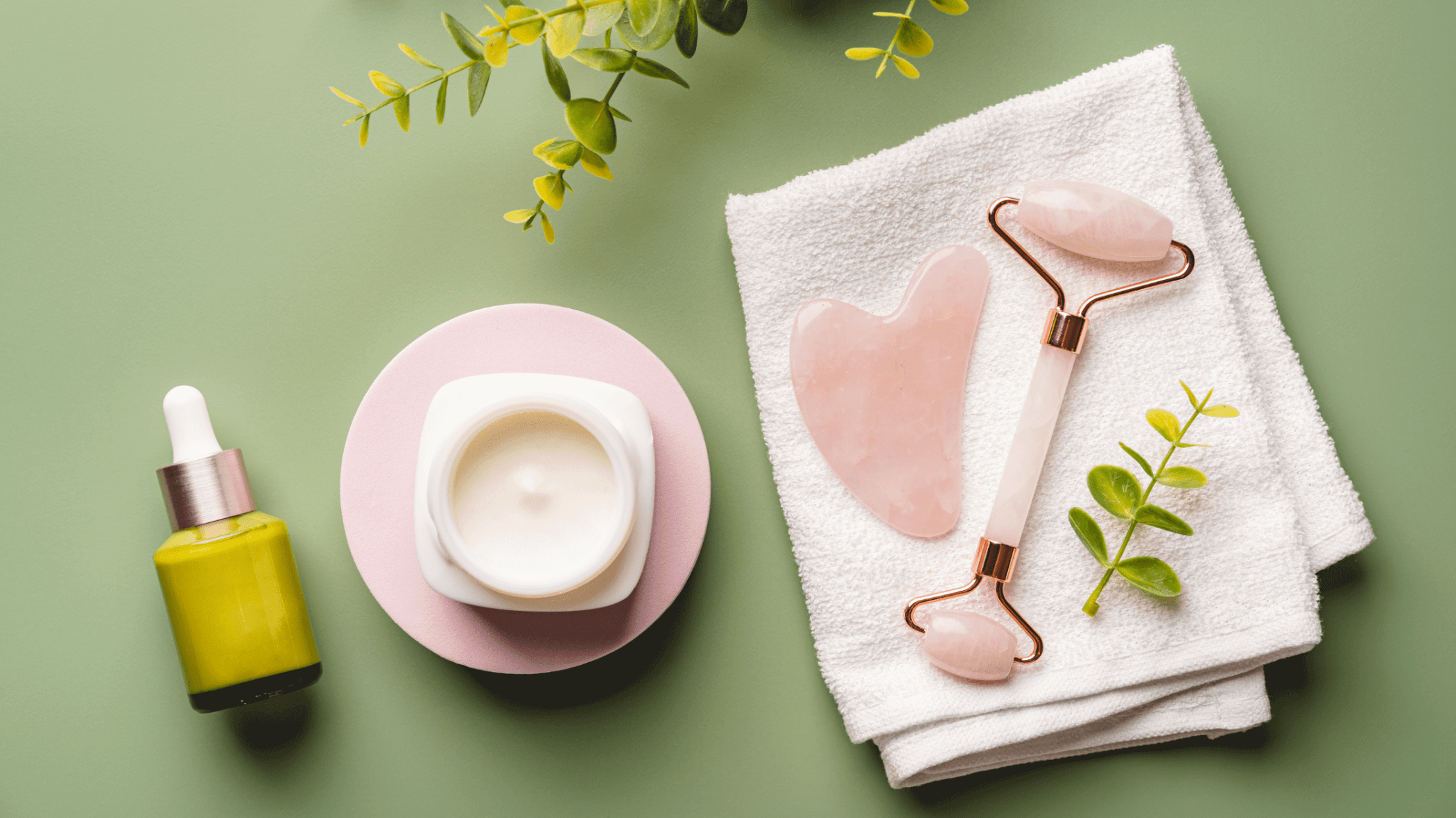 Spa and skincare products arranged on a sage green background including a golden facial oil, rose quartz facial rollers, a cream moisturizer, white towels, and fresh green botanical sprigs.