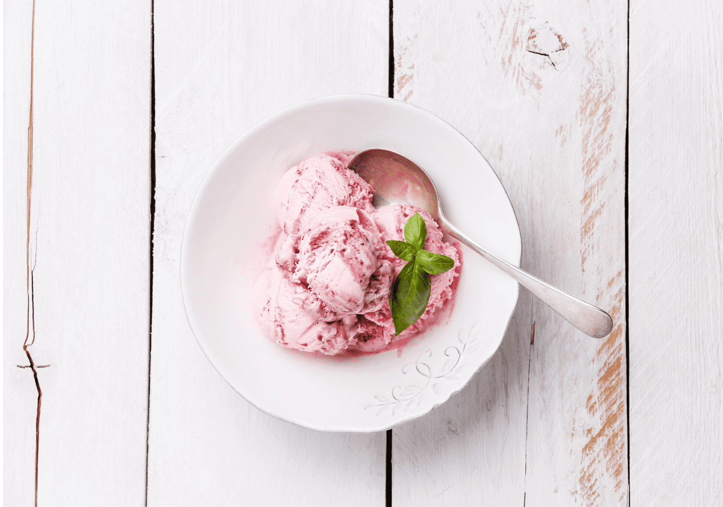 Three scoops of pink ice cream garnished with fresh mint leaves in a white bowl with a spoon on a whitewashed wooden surface.