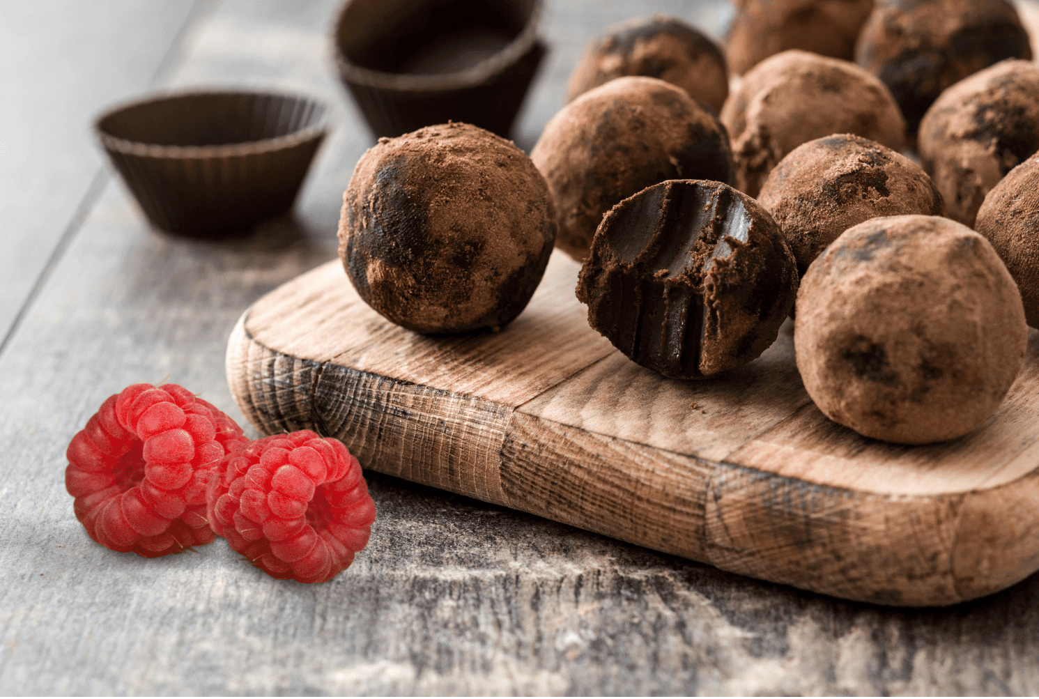 Chocolate truffles dusted with cocoa powder arranged on a wooden board with fresh red raspberries displayed on a gray surface.