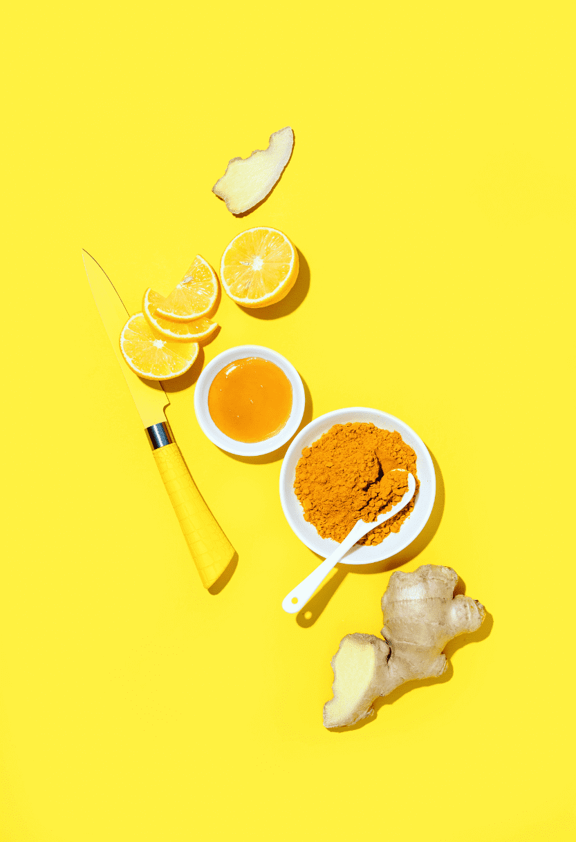 Turmeric powder, fresh ginger root, lemon slices, ginger root pieces, honey, and a yellow measuring spoon arranged on a yellow background.