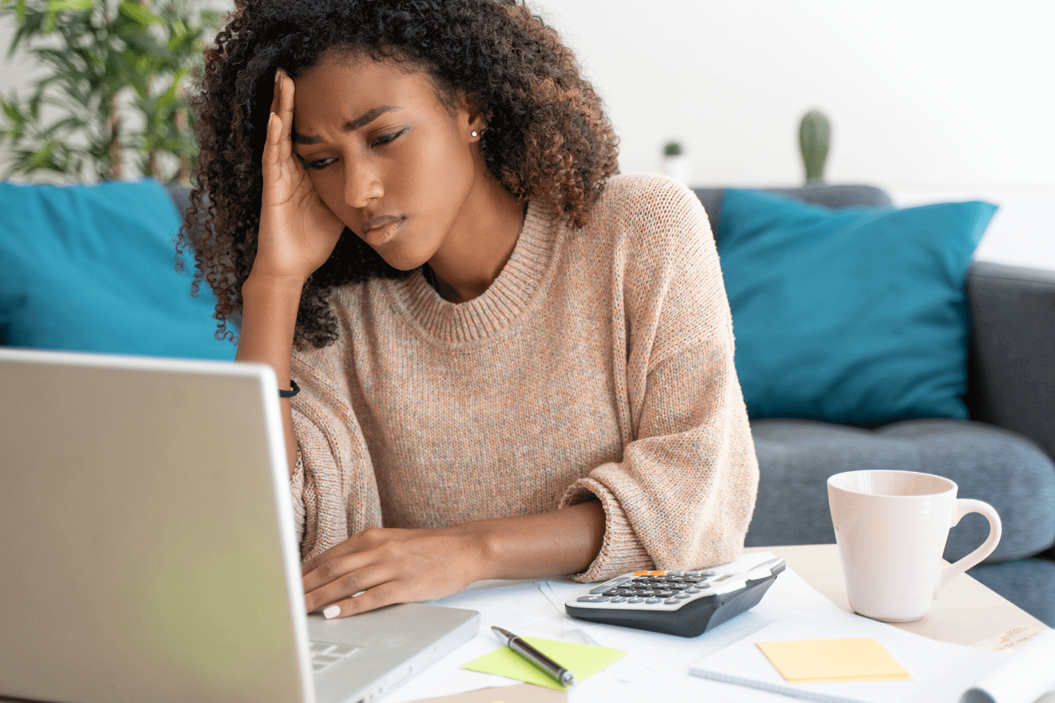 Woman with curly hair experiencing stress or fatigue while working at a laptop surrounded by financial documents and a calculator.