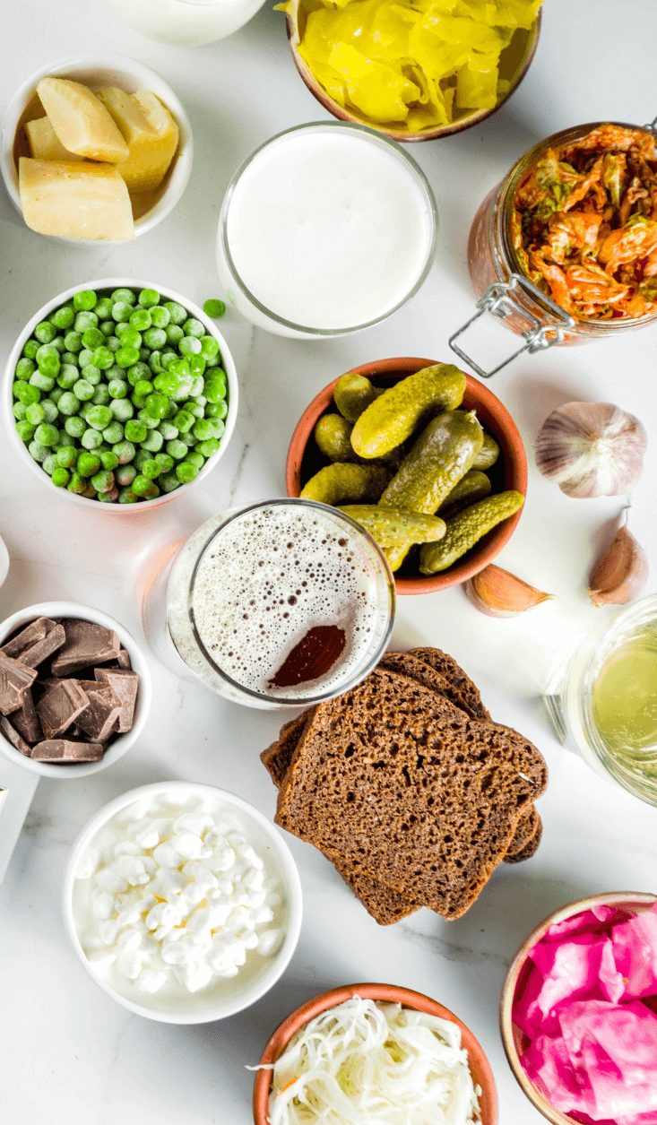 Overhead flat lay of fresh ingredients arranged on a white surface including pickles, peas, bread, beer, mayonnaise, pickled vegetables, garlic, oil, and other components for meal preparation.
