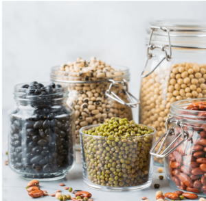 Glass jars filled with various legumes and beans including black beans, chickpeas, green lentils, and red kidney beans arranged on a white surface.