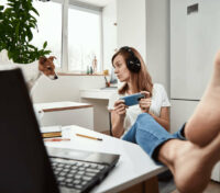 Woman wearing headphones holds a mobile device while working at a desk in a bright home office with a dog nearby.