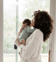 A woman holding a young child looks out a large window toward natural outdoor scenery.