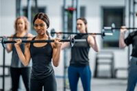 A woman performs a barbell shoulder press in a gym while other participants exercise in the background.