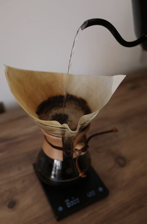 Water is being poured from a kettle into a coffee filter filled with ground coffee in a glass brewer positioned on a scale.