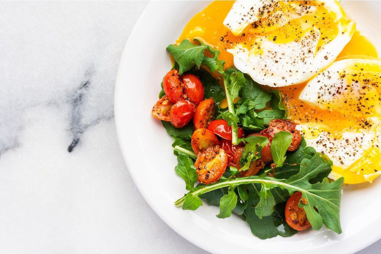 A plate contains two poached eggs topped with black pepper, served alongside a salad of arugula and roasted cherry tomatoes.