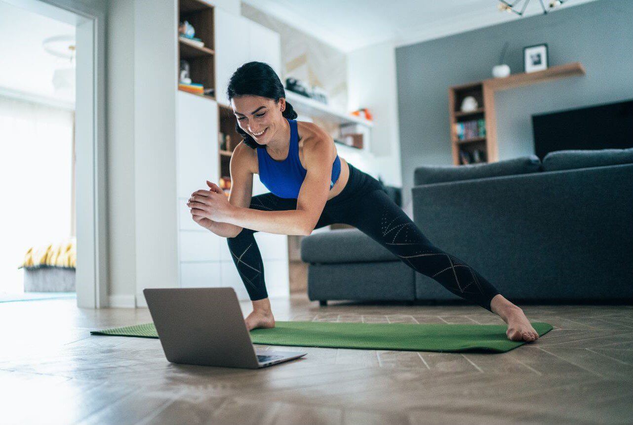 A woman in athletic wear performs a plank exercise on a green mat while following an online workout video on her laptop in her living room.