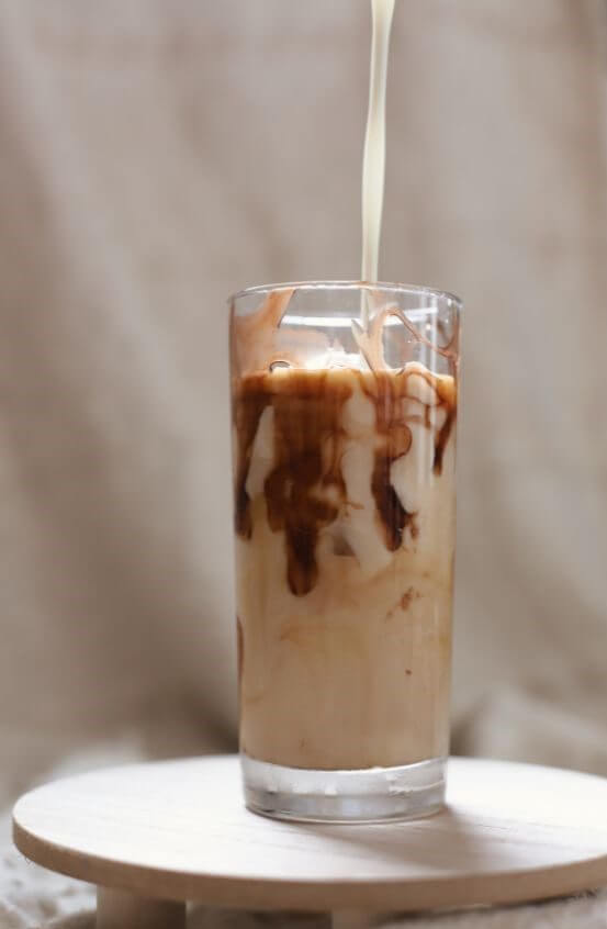 A glass filled with creamy iced coffee is being poured with milk, creating swirls of color.