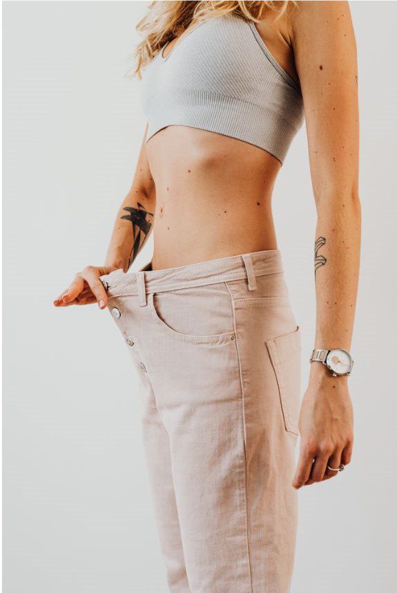 A woman with tattoos in a bra top stands confidently against a white wall, showcasing her weight loss journey.
