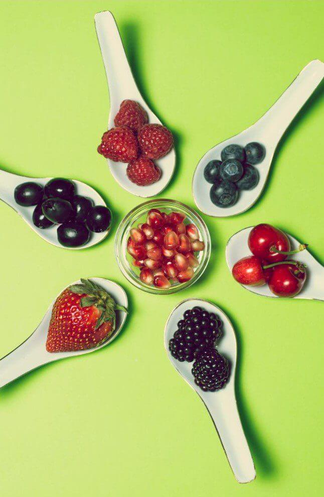 Various fruits, including raspberries, blueberries, cherries, black olives, a strawberry, and blackberries, are arranged in white spoons surrounding a small bowl of pomegranate seeds on a green background.