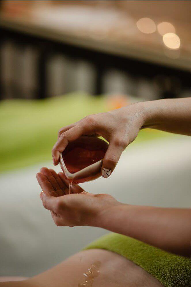 A person pours oil from a small bowl into their hands while preparing for a massage on a green towel.