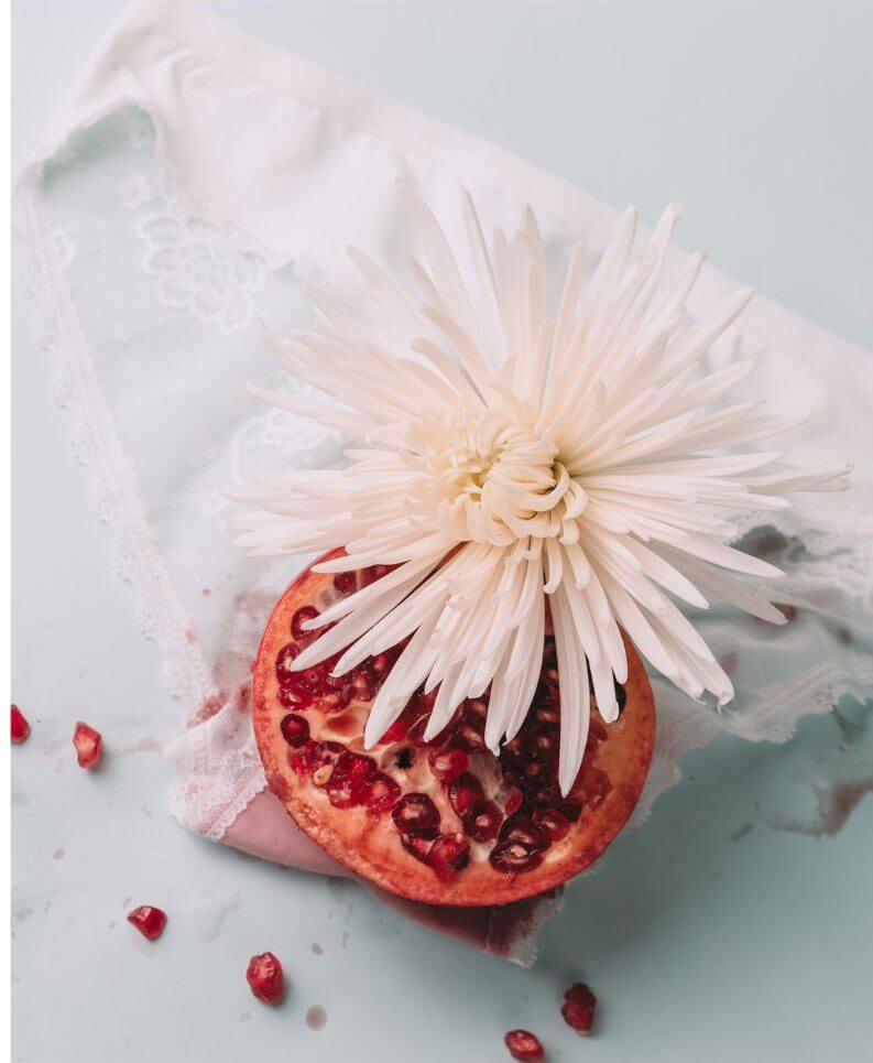 A pomegranate half and a white flower are placed on a light blue surface, with some pomegranate seeds scattered around.