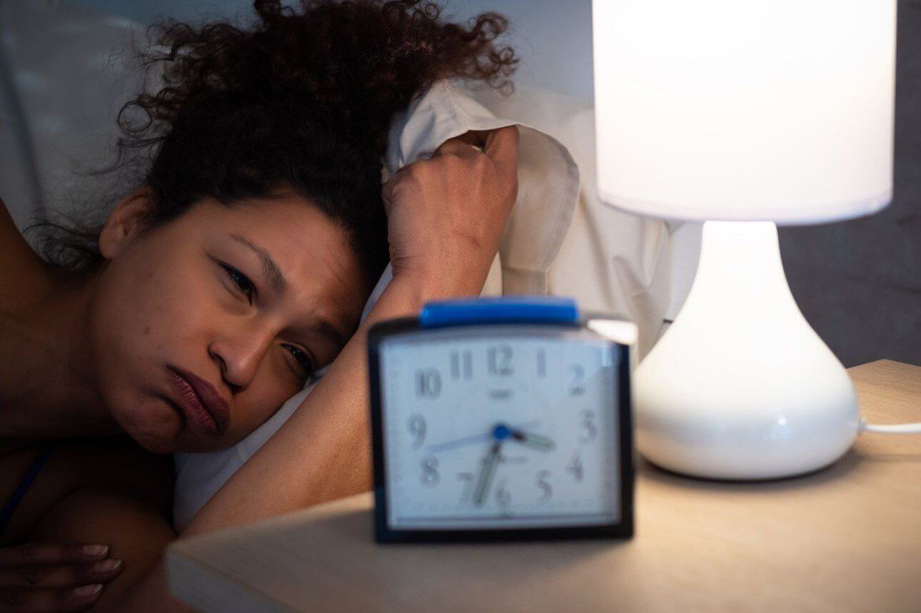 A woman with curly hair lies in bed, resting her head on her arm while looking at an alarm clock on the bedside table.