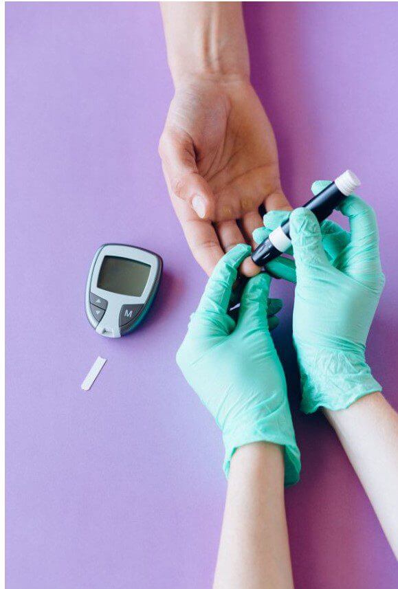 A person wearing green gloves is using a glucose meter on the hand of another person against a purple background.