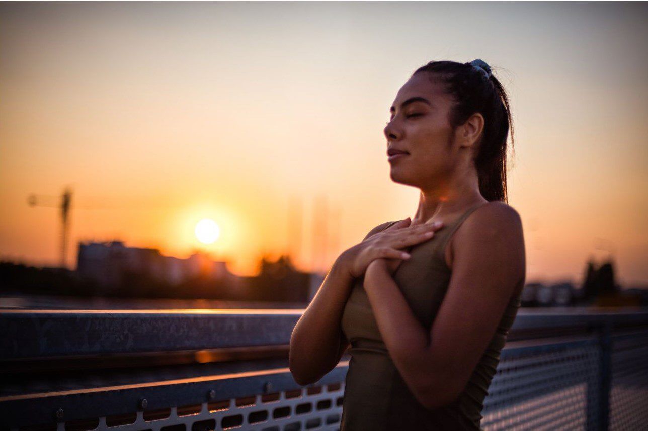 A woman in a green tank top stands with her hands over her chest, facing a sunset while closing her eyes.