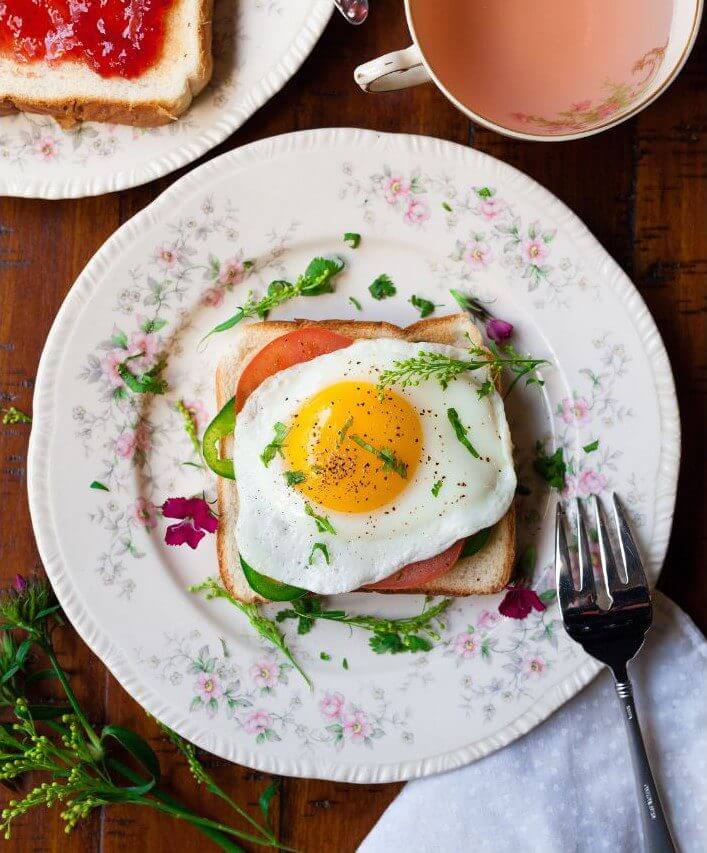 A slice of toast topped with a fried egg, tomato, and green pepper is served on a decorative plate with fresh herbs and a fork beside it.