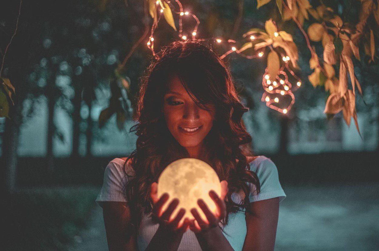 A woman with long, wavy hair smiles while holding a glowing, round object that resembles the moon, surrounded by greenery and soft, warm lights.