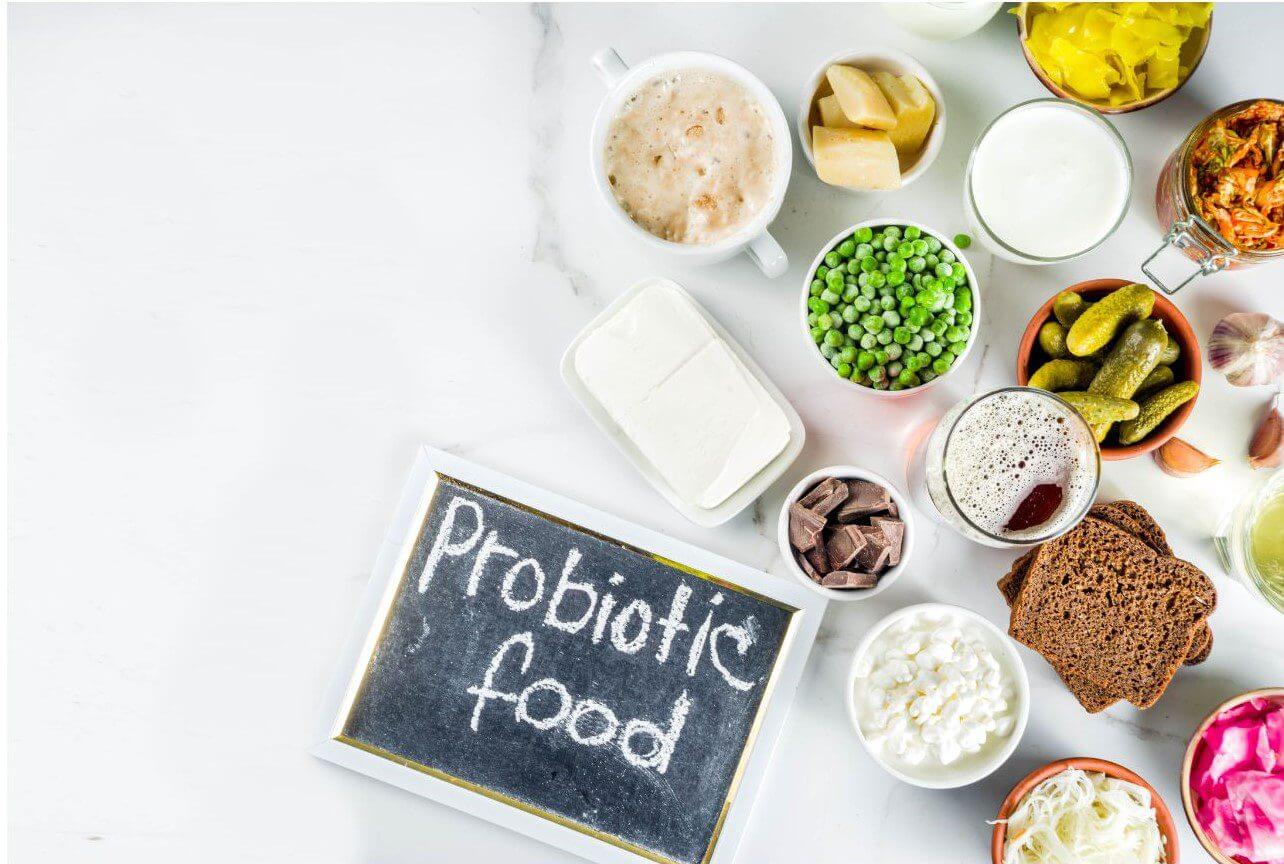 Various probiotic foods, including fermented vegetables, yogurt, and grains, are arranged on a marble surface alongside a chalkboard label reading 'Probiotic food.'