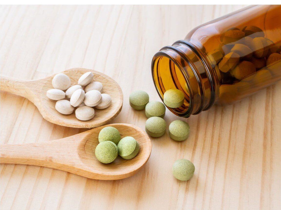 Brown glass bottle tipped on its side with assorted tablets spilling out onto a wooden surface, with white tablets on one wooden spoon and green tablets on another.