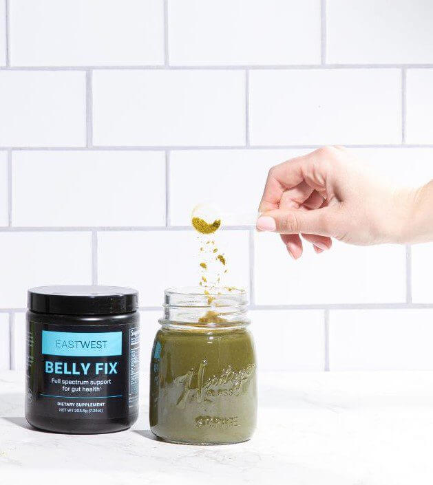 A glass jar filled with green juice stands next to a hand holding a metal spoon.
