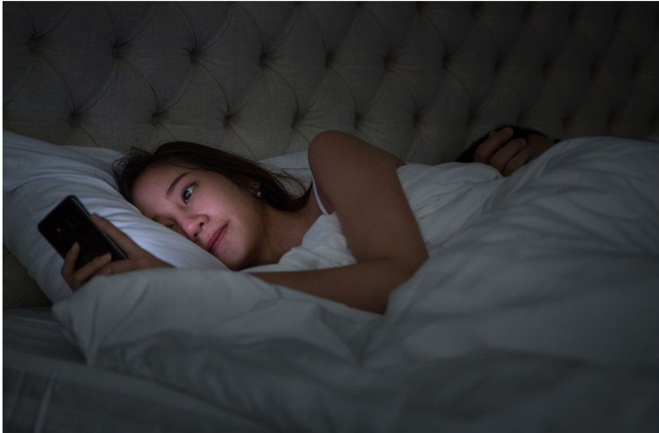 A woman is lying in bed, looking at her smartphone while resting on white pillows and bedding.