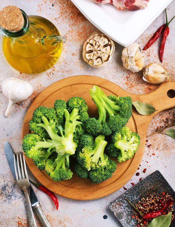 A wooden cutting board displays fresh green broccoli alongside garlic, spices, and a bottle of olive oil.
