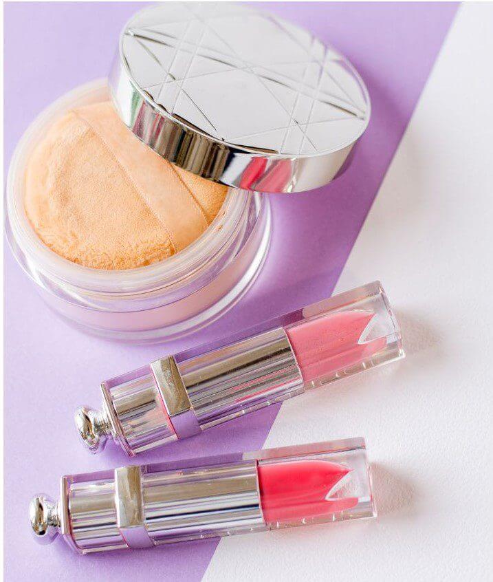 A compact container of loose powder and two tubes of pink lip gloss are placed on a purple background.