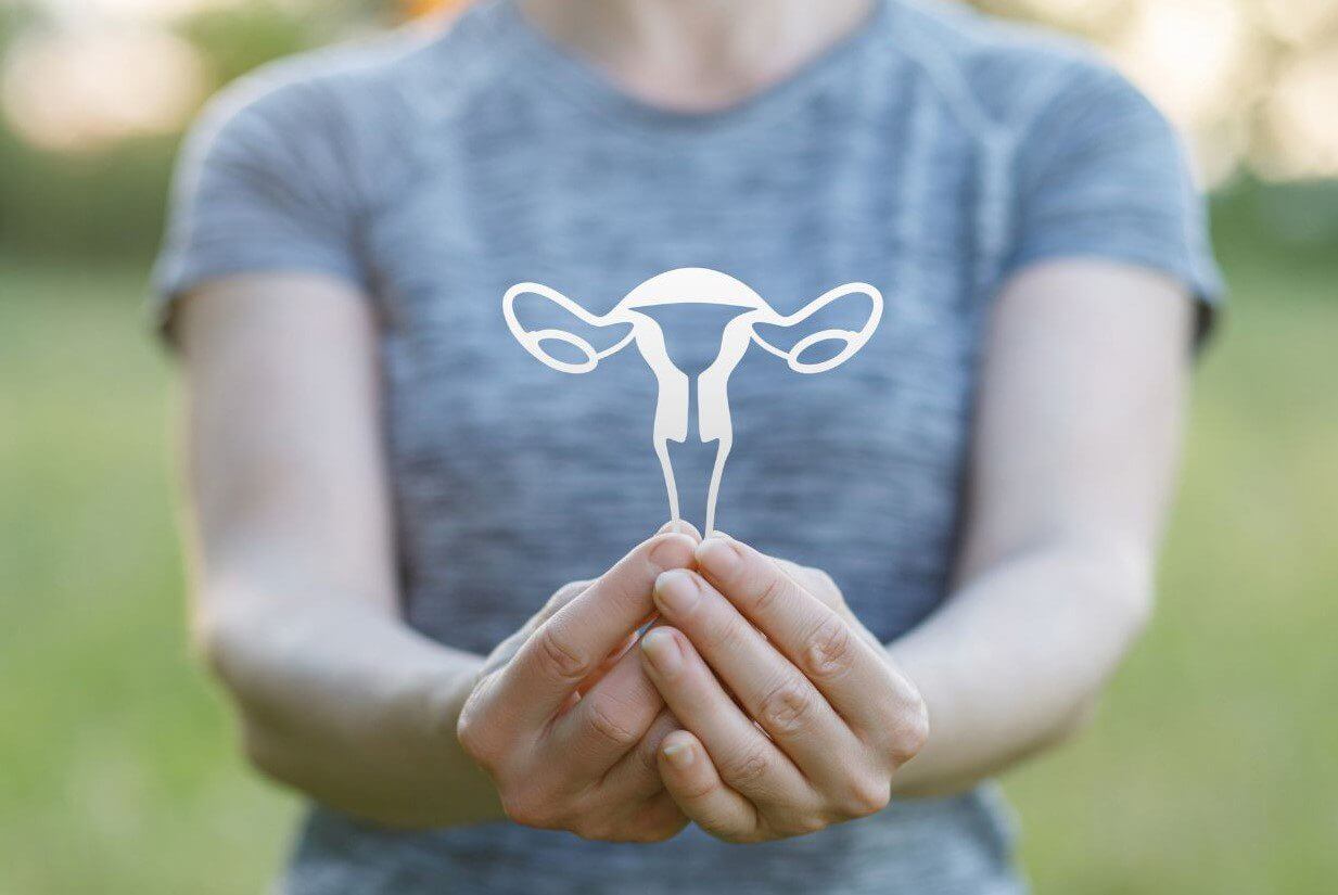 A person in a gray athletic shirt holds a white cut-out representation of a female reproductive organ in a green outdoor setting.