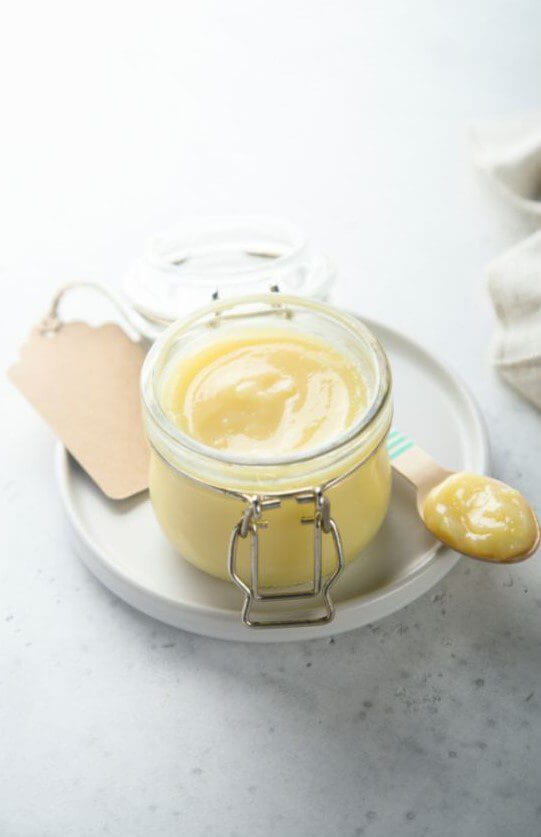 A clear jar filled with yellow creamy substance sits on a round plate alongside a spoon with a small amount of the substance on it.