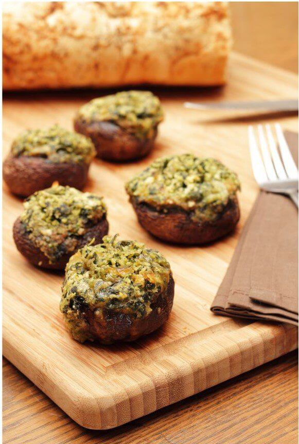 Stuffed mushrooms with a green topping are arranged on a wooden cutting board beside a piece of bread and a brown napkin.