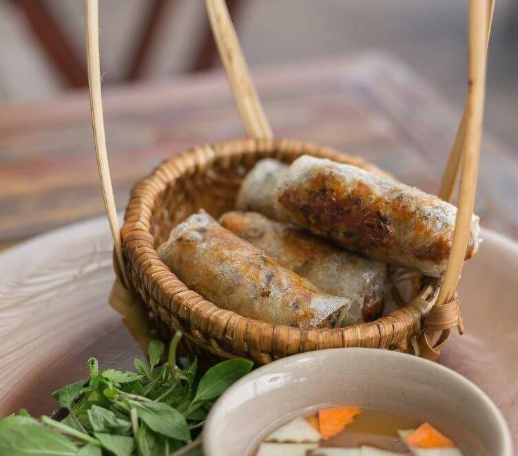 A woven basket contains several spring rolls alongside a small bowl of dipping sauce and fresh herbs.