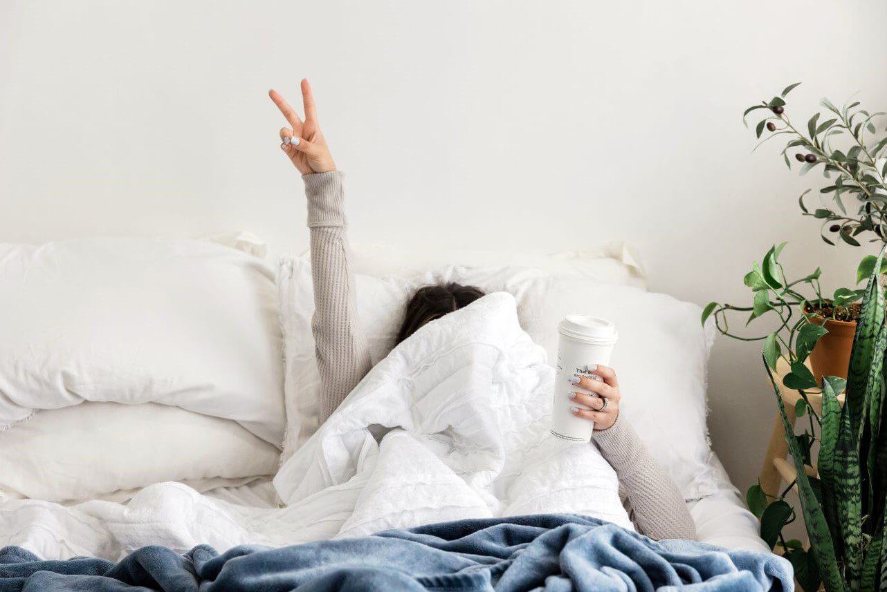 A person wrapped in blankets raises one arm in a peace sign while holding a coffee cup in the other hand.