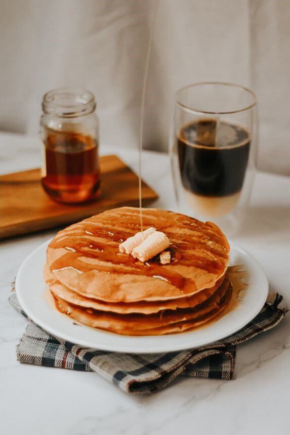 Pancakes topped with butter are served on a white plate with syrup drizzling over them, accompanied by a glass of dark beverage and a jar of syrup.