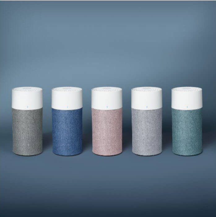 Five cylindrical speakers with fabric textures in gray, blue, pink, light gray, and teal are arranged in a row against a blue background.